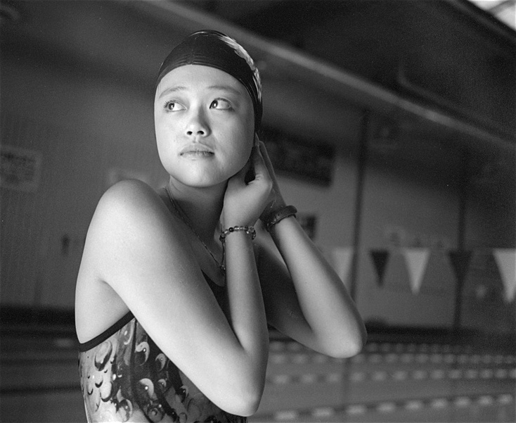 Black-and-white photograph by Barbara Beirne. At an Olympic pool, Lili Shek, a young Chinese woman, is wearing a bathing suit and adjusting her swim cap. She's looking up and to the left of the frame.